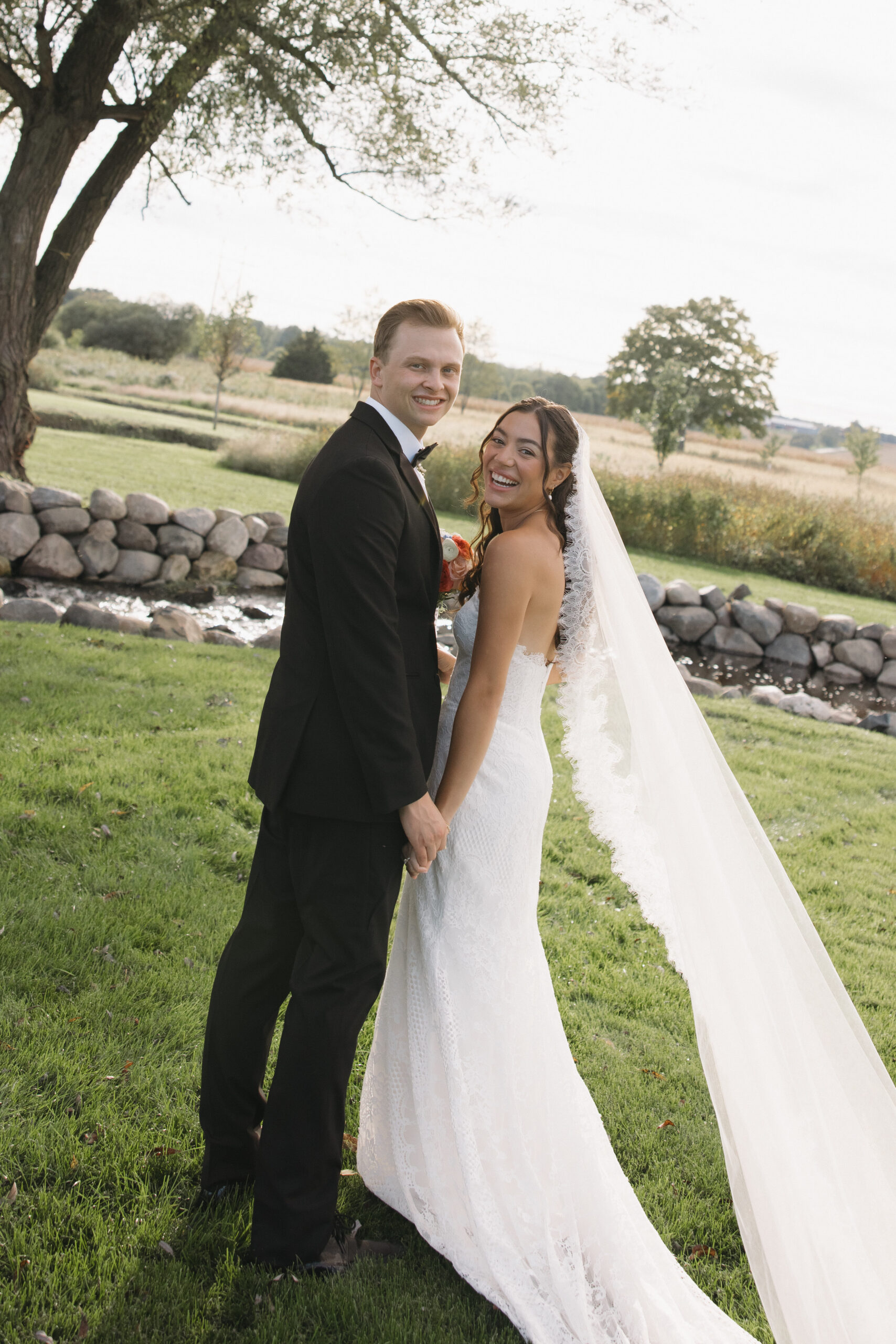 Bride and groom holding hands under a willow tree by the stone lined pond at Covered Bridge Estate wedding and event venue